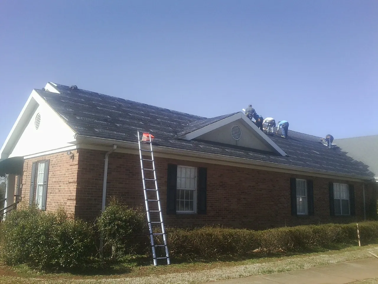 Crew tearing shingles off a brick ranch roof with a ladder set — Emergency Roof Repair work by our crew in Spring Glen